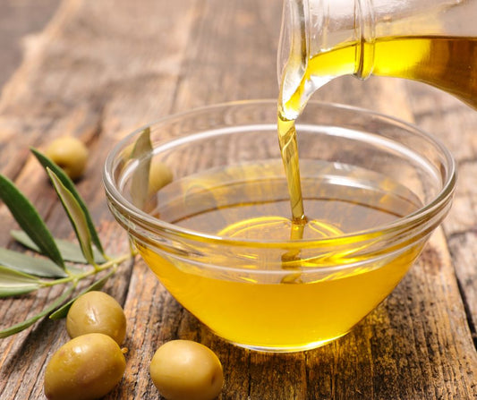 Olive oil being poured into a glass bowl on a wood table with olive leaves and fruit nearby.