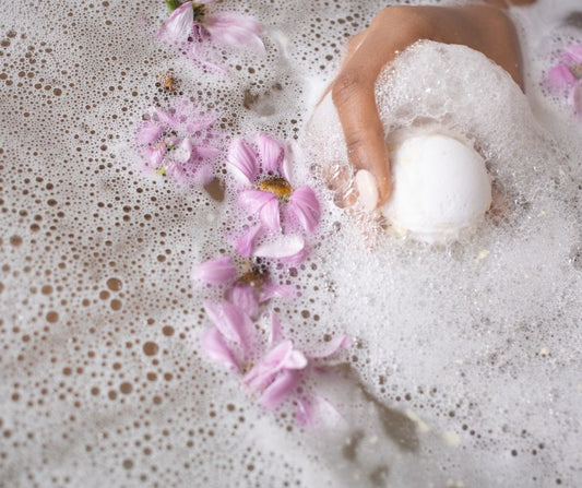 A woman's hand holding a fizzy bath bomb in a tub next to pink flowers.
